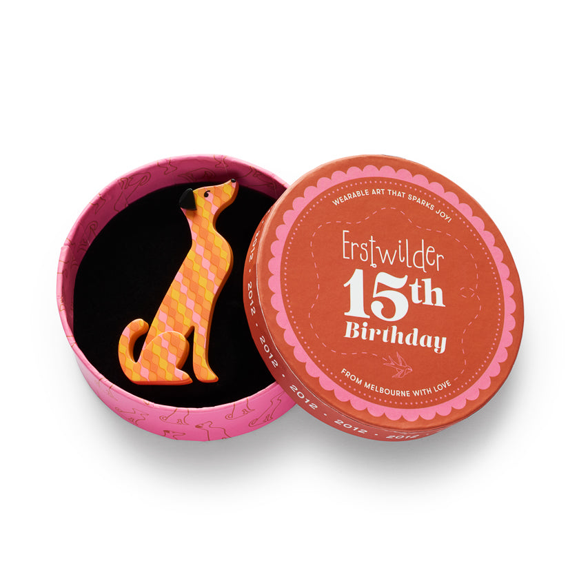 An open round pink gift tin with a dog-shaped brooch inside, alongside the lid which reads 'Erstwilder 15th Birthday' on an orange label, set against a plain white background.