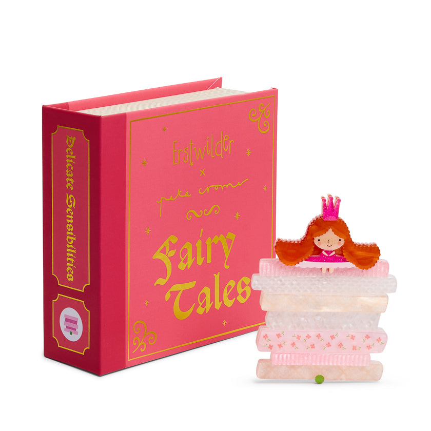 Pink book titled 'Fairy Tales' with a small figurine of a girl in a princess dress on top of a stack of mattresses and a pea.