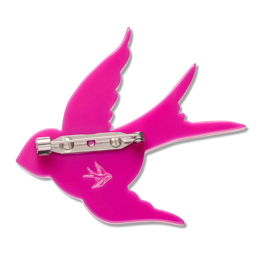 A bright pink enamel pin shaped like a flying swallow, featuring a silver-tone metal pin clasp on the back and a small silver swallow design printed on the front, set against a plain white background.