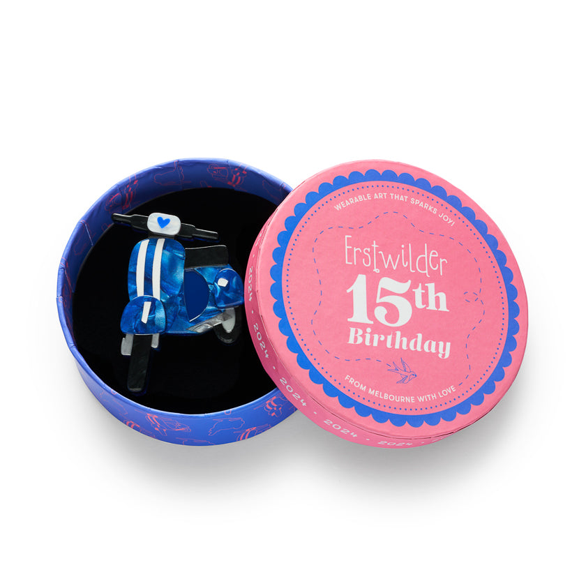 A small blue toy vespa brooch inside an open round presentation box, with the pink and blue lid placed beside it on a white background. The lid reads 'Erstwilder 15th Birthday' and 'FROM MELBOURNE WITH LOVE'.